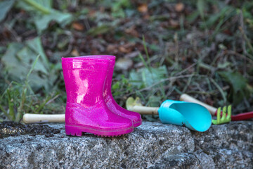 Set of gardening tools displayed side by side, outdoors on a rock and vegetation as a background.  Child's pink wellies in focus.