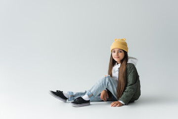 full length of adorable girl in trendy autumnal outfit and beanie hat sitting on grey.