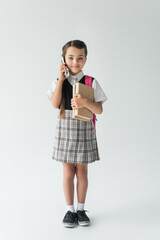 full length of smiling schoolgirl talking on smartphone and holding books on grey.