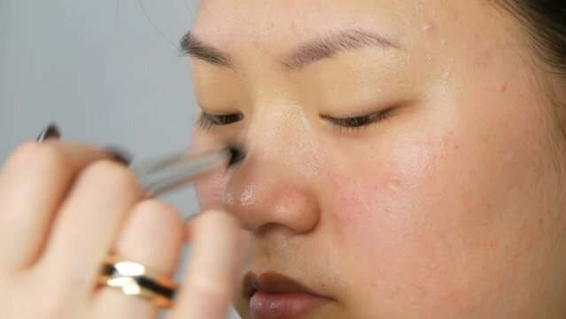 Professional makeup artist applies foundation concealer or highlighter to Asian Korean model's face with special brush
