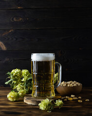 Transparent high mug with light beer on a wooden table. Hop cones in the background. Wooden bowl with peanuts on the table. Dark brown wooden background