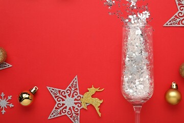 Flat lay composition with shiny confetti spilled out of champagne glass near Christmas decorations on red background, space for text