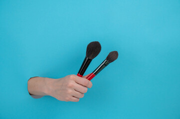 A woman is holding two large brushes for cosmetics. Woman's hand sticking out of blue paper background. 