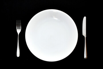 Stainless steel knife and fork and empty white dinner plate on a plain back background. No people.