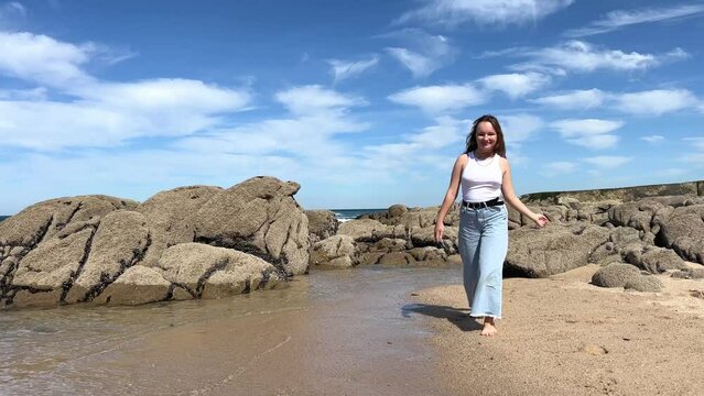a teenage girl in white t-shirt and jeans walks along a stone beach in Portugal on atlantic ocean she is happy spreading her arms to the sides and looking at the water good for traveling and relaxing.