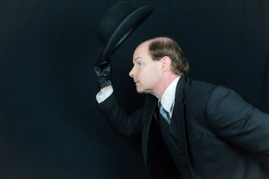 Portrait of British Businessman in Black Suit and Leather Gloves Tipping Bowler Hat and Bowing in Polite Greeting. Classic English Gentleman or British Butler.