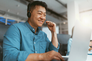 Smiling asian businessman in headphones talking online with client sitting in office