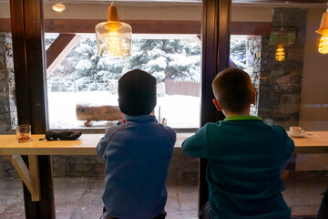 two children with their backs to each other sipping hot chocolate in a ski resort bar while gazing out the window at the snow-covered landscape