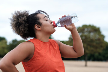 Sport curvy woman is drinking a bottle of water in a park.