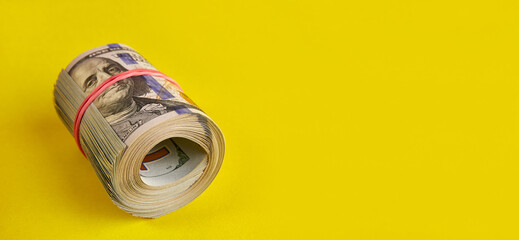 Many hundred dollar bills rolled up with a red rubber band on a yellow background.