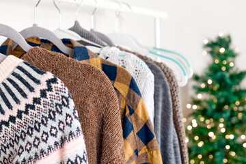 Knitted sweaters on hangers on clothes rack in the shop with blurred christmas tree background. Slow Fashion. Conscious consumption. Black friday. Winter sale.