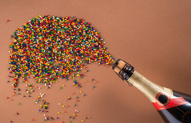 Bottle of champagne with splash of many small colored balls on brown studio background. Bright multicolored round particles of red white, blue, green and yellow with workspace. Wallpaper for holiday.
