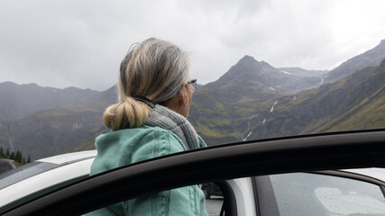 A gray-haired woman in a warm sweater stands near a car in an alpine valley and looks at the mountains.
