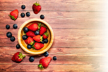 A wooden bowl with strawberries and blueberries on a wooden table. Top view with space for text and flare effect.