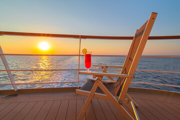 Vue d'une chaise longue avec un cocktail sur le pont d'un navire de croisière au coucher de soleil dans le sillage du navire de croisière. Vue depuis la poupe du navire.	