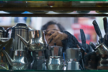 Woman picks up a small silver kettle from a glass shelf inside a store in an Arabic market. Focus is on hand, perspective from outside the shop.