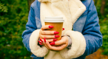 Paper cup with coffee in hands of unrecognizable woman, standing in forest. Body part of female in warm clothes with bright manicure holding hot drink.