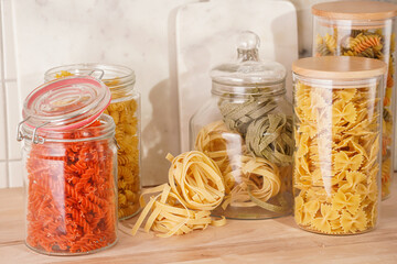 Variety of dry italian pasta in glass jars on kitchen cabinet with wooden top: fusilli, tagliatelle, farfalle, spaghetti