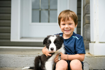 Happy boy hugging black and white border collie in front of house door - obrazy, fototapety, plakaty