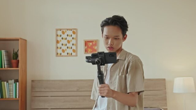 Teenage Asian boy using handheld gimbal stabilizer and smartphone while filming lifestyle vlog at home