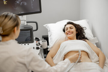 Happy adult woman during an ultrasound examination of the abdominal cavity at modern medical office. Concept of women's health and examination during pregnancy - obrazy, fototapety, plakaty