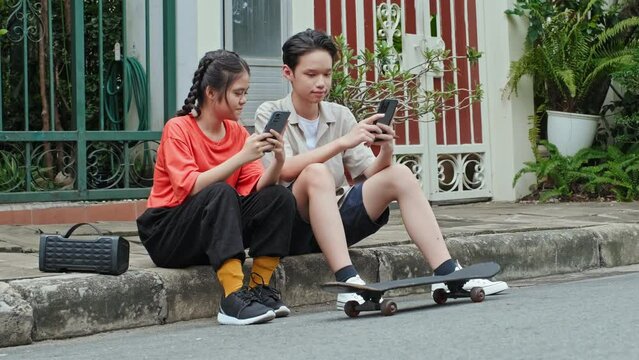 Asian boy and girl sitting together on street curb beside skateboard and portable speaker, browsing the web on smartphones and chatting