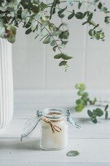White wax candle in a glass jar decorated with twine on a white wooden background with eucalyptus sprigs. Minimalism. High key.