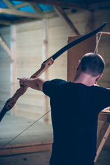 A man in a shooting gallery holds a bow in his hands and aims. The bowstring is stretched from the back.