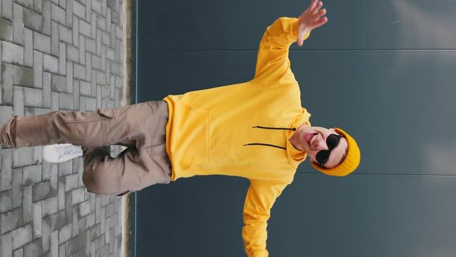 Young Man caucasian dancing moving his hands with his legs with making funny faces wearing yellow hoodie, beanie hat sunglasses close up against blue sky. Younger generation. Lifestyle. Vertical video