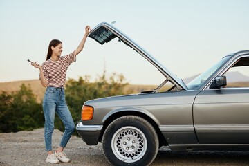 A woman stands outside a broken-down, dangerous old car with a wrench on the road traveling alone