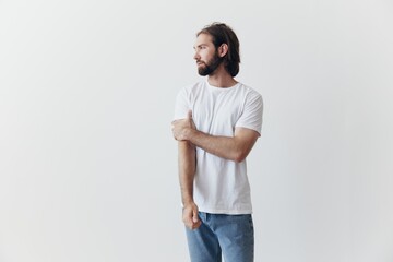 A man with a beard and long hair in a white T-shirt and blue jeans stands against a white wall...