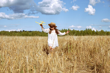 A little girl in a white dress and a straw hat plays with a toy plane in a rye field in