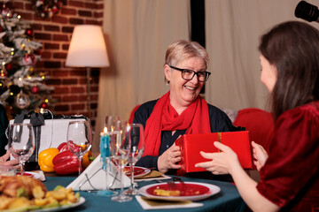 Family exchanging christmas gifts at table, celebrating winter holiday, having festive dinner, eating traditional dishes. Xmas celebration with parents, daughter giving mom present box