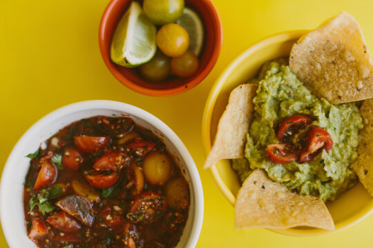 Overhead view of salsa and guacamole served with ingredients on yellow background
