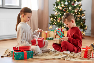 christmas, winter holidays and childhood concept - happy girl and boy in pajamas opening gifts sitting on floor in front of each other at home