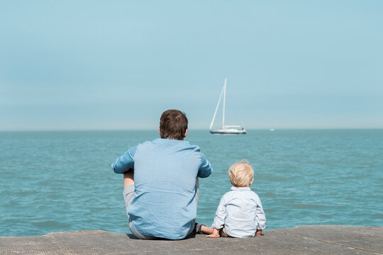 Rear view of father with son sitting at beach against blue sky during sunny day