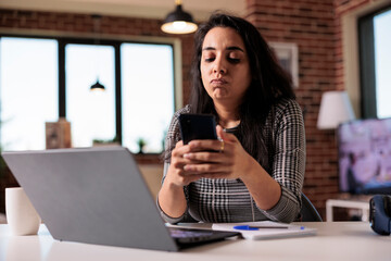 Young adult browsing internet on smartphone, using social media app to text messages on work break at home. Working on remote job with laptop and mobile phone, searching online information.