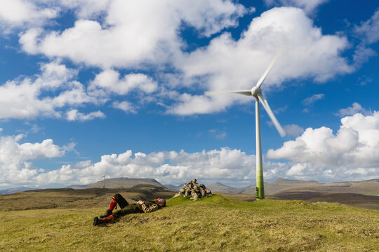 Man lying on grassy field by windmill against cloudy sky