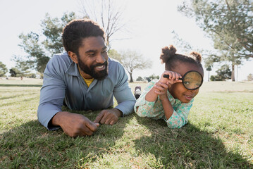 Portrait of daughter playing with magnifying glass while lying by father on grassy field at park during sunny day