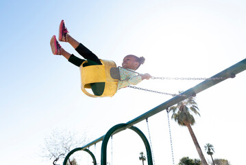 Low angle view of playful girl swinging against clear sky at playground during sunny day