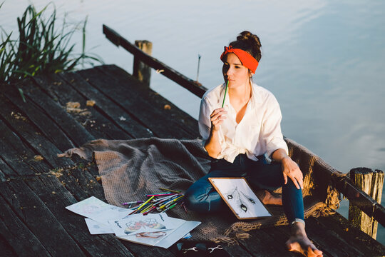 High angle view of thoughtful woman drawing while sitting on pier over lake during sunset
