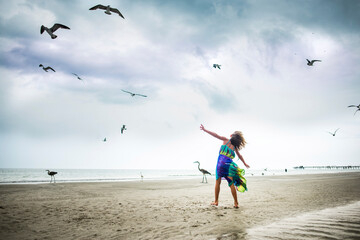 Full length of playful girl with arm outstretched standing at beach while birds flying against cloudy sky