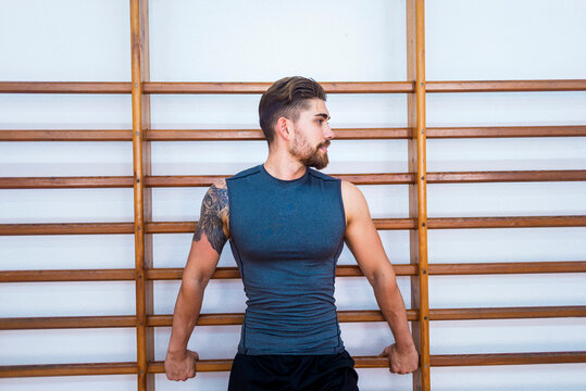 Muscular tattooed athlete looking away while standing against wall bars in gym