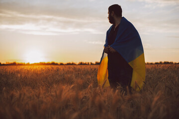 Thoughtful man with Ukrainian Flag standing amidst crops on field against sky during sunset