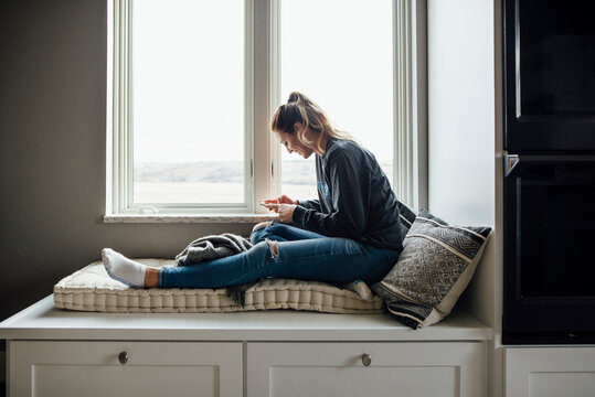Side view of woman using mobile phone while sitting on alcove window seat at home