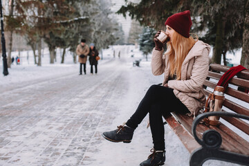 Woman drinking coffee while sitting on bench against trees at park during winter