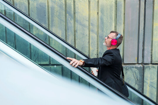 Side view of businessman listening music while standing on escalator against wall