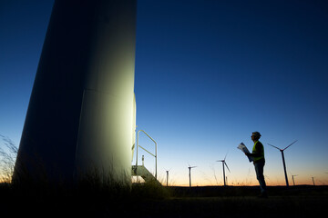 Side view of silhouette engineer looking at windmill while standing on field against clear blue sky during sunset
