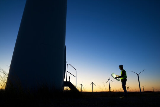 Side View Of Silhouette Male Engineer Looking At Paper While Standing By Windmills On Field Against Clear Sky During Sunset