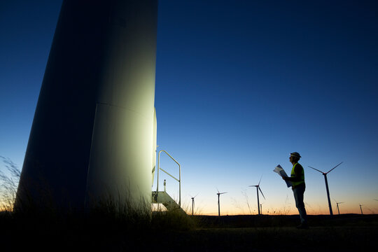 Side view of silhouette engineer looking at windmill while standing on field against clear blue sky during sunset